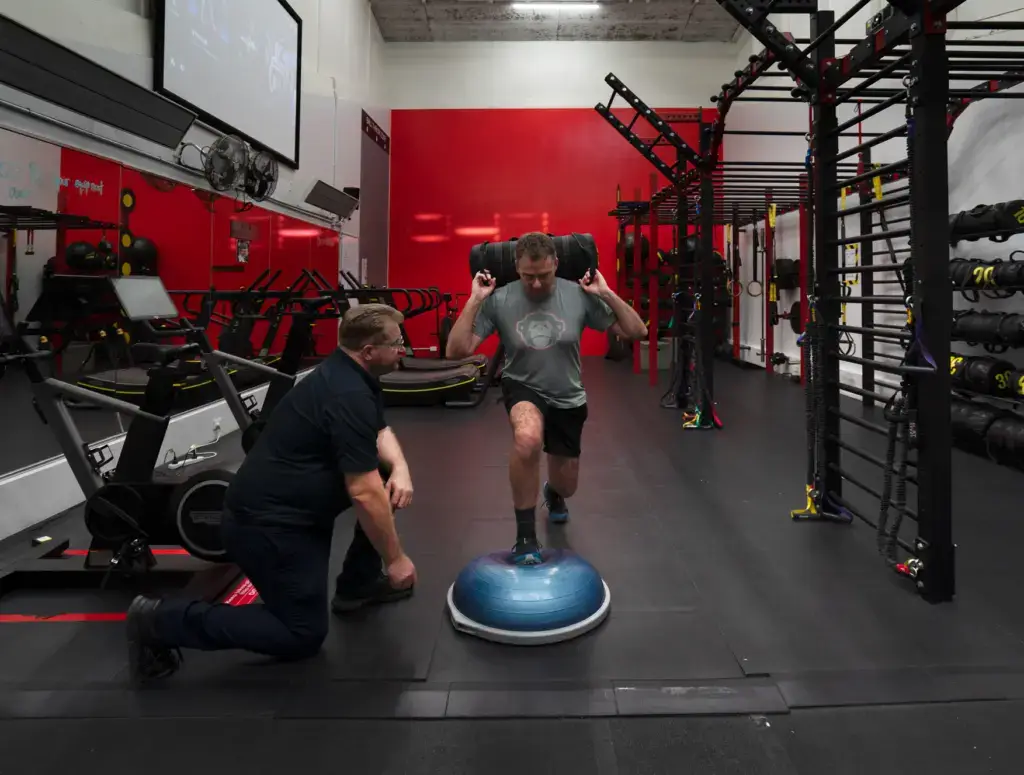 Man balancing on BOSU with trainer spotting